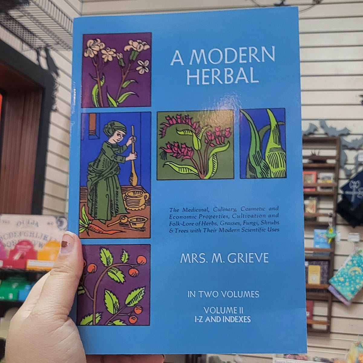 A hand holding a blue book titled 'A Modern Herbal' in a home interior setting with wooden paneling and shelves in the background.