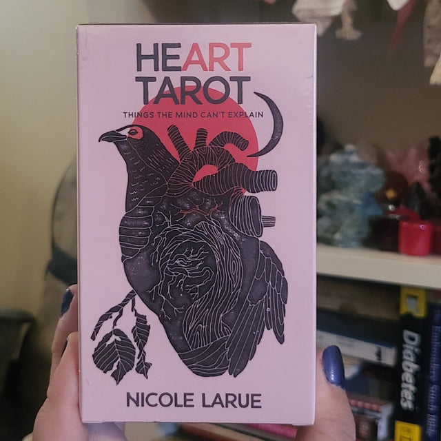 A hand holds a pink tarot card deck box titled 'Heart Tarot' by Nicole Larue, featuring a black anatomical heart illustration with a red heart shape. The background shows a cozy indoor setting with books, a shelf, and sunflowers.