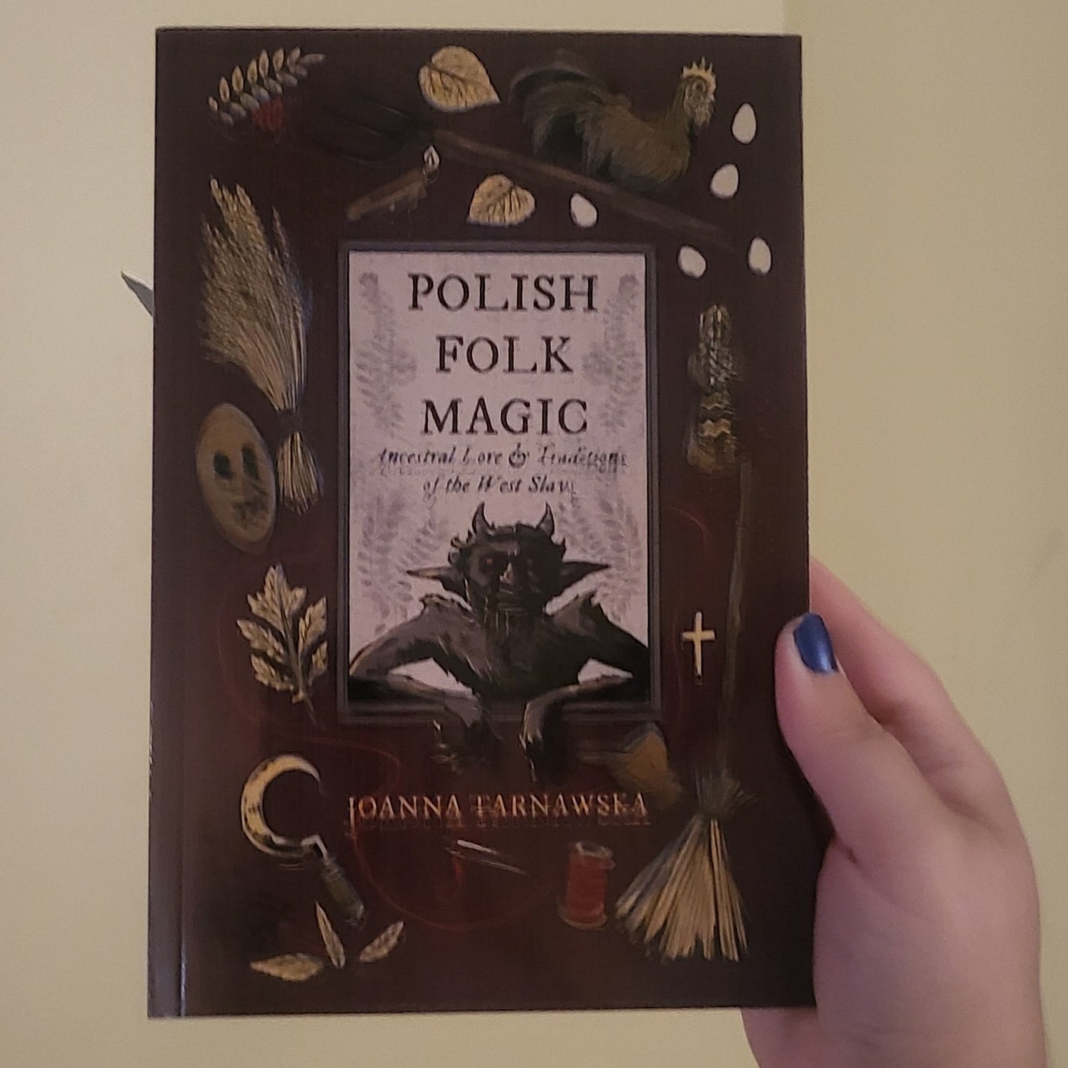 A hand holds a book titled 'Polish Folk Magic' against a plain beige wall. The book cover is dark brown with occult illustrations including a cat, pentacle, and candles.