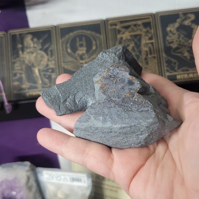 Hand holding a gray stone against a blurred background with various items.
