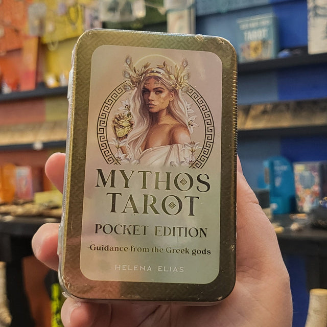Hand holding a tin labeled 'Mythos Tarot' against a blurred store background