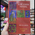 A hand holding a book titled 'A Modern Herbal' by Mrs. M.