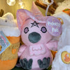 Plush crow doll named "Alice Crow-ley", viewed from the front. Doll is overall pink except for violet embroidered eyes, grey plush beak and feet, and black embroidered details for pentagram and eyeliner. It has pink plush wings and feather beard with embroidered pink detail. It also has a pink witch's hat with white star print with a violet hat band.