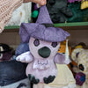 Plush crow doll named "Marie Corveau", viewed from the front. Doll is overall violet except for purple embroidered eyes, black plush beak and feet, and black embroidered details for pentagram and eyeliner. It has violet plush wings and feather beard with embroidered purple detail. It also has a purple witch's hat with dark purple floral print and black hat band.