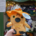 Plush crow doll named "Maggie Pie", viewed from the front. Doll is overall orange except for black embroidered eyes, black plush beak and feet, and black embroidered details for pentagram. It has orange plush wings and feather beard with embroidered yellow detail. It also has a candy corn-coloured witch's hat.