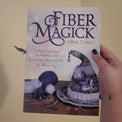 A hand with dark nail polish holds up a book titled 'Fiber Magick' by Opal Luna against a beige wall.