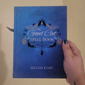 A hand holding a blue hardcover book titled 'The Good Cat Spell Book' by Gillian Kemp against a plain beige wall.