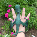 Hand wearing a green dragon-shaped glove with pink flowers and greenery in the background