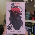 A hand holds a pink tarot card deck box titled 'Heart Tarot' by Nicole Larue, featuring a black anatomical heart illustration with a red heart shape. The background shows a cozy indoor setting with books, a shelf, and sunflowers.
