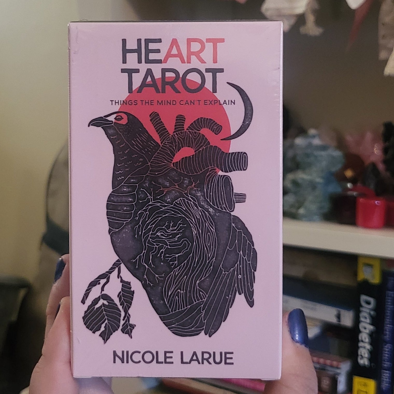 A hand holds a pink tarot card deck box titled 'Heart Tarot' by Nicole Larue, featuring a black anatomical heart illustration with a red heart shape. The background shows a cozy indoor setting with books, a shelf, and sunflowers.