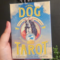 A hand holding a boxed tarot card deck titled 'The Dog Tarot' against a dark background with decorative flowers and dried lavender.