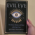 A hand holding a book titled 'The Evil Eye' by Antonio Pagliarulo against a plain beige wall. The book cover is black with a gold and blue eye illustration and gold decorative borders.