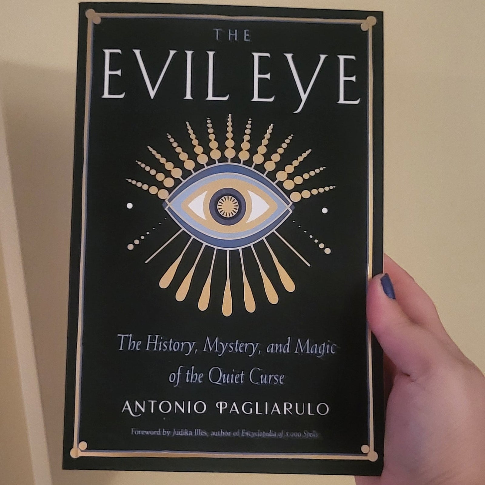 A hand holding a book titled 'The Evil Eye' by Antonio Pagliarulo against a plain beige wall. The book cover is black with a gold and blue eye illustration and gold decorative borders.