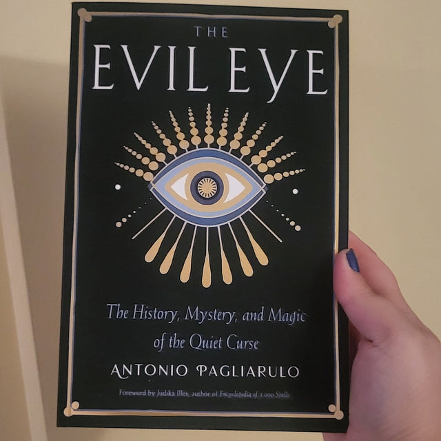 A hand holding a book titled 'The Evil Eye' by Antonio Pagliarulo against a plain beige wall. The book cover is black with a gold and blue eye illustration and gold decorative borders.