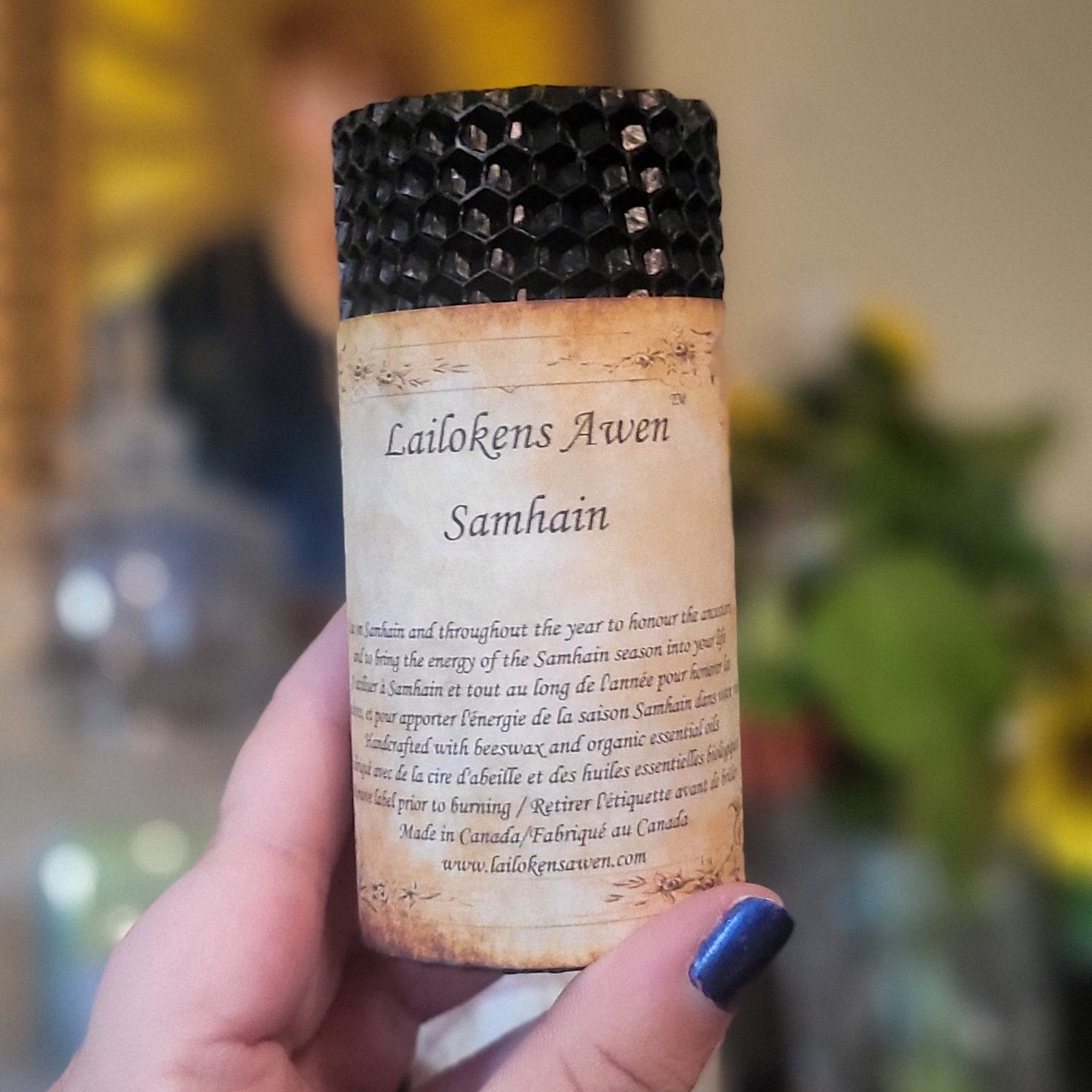 A hand with blue nail polish holds a small cylindrical candle with black honeycomb beeswax and a parchment-style label reading 'Samhain'. The background is a warm, blurred indoor setting with a framed sun illustration and a plant.