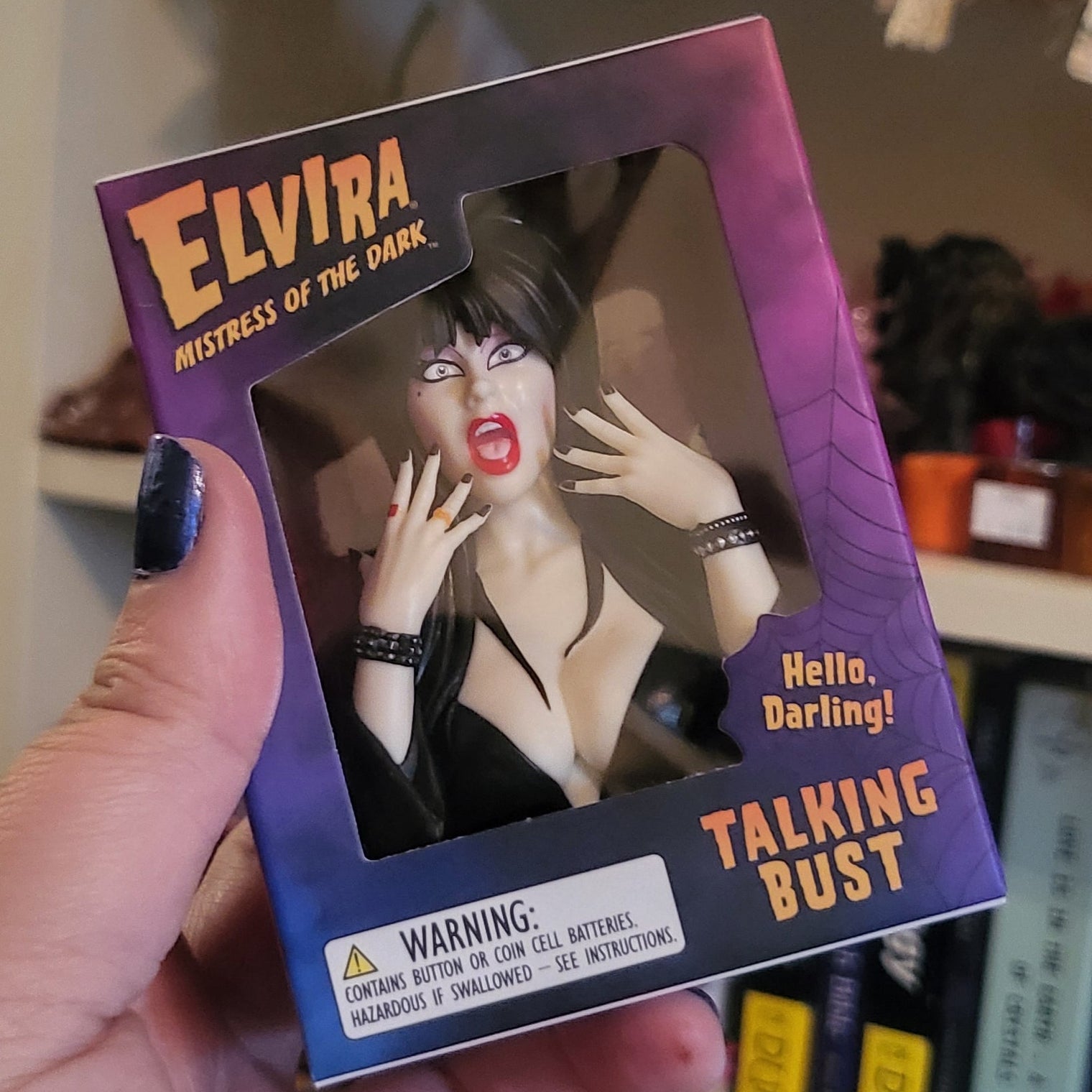 A hand holds a collectible figure of Elvira Mistress of the Dark in its original packaging, which features a purple and black box labeled 'Elvira Talking Bust'. The background shows a cluttered indoor shelf environment.